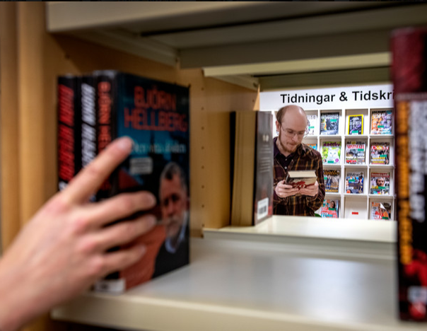 En hand ställer in böcker i en bokhylla. Bakom skymtas en man som står och läser framför hyllan märkt Tidningar & tidskrifter.